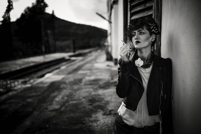 Woman in a leather jacket holding a cigarette while leaning against a wall at an outdoor train platform
