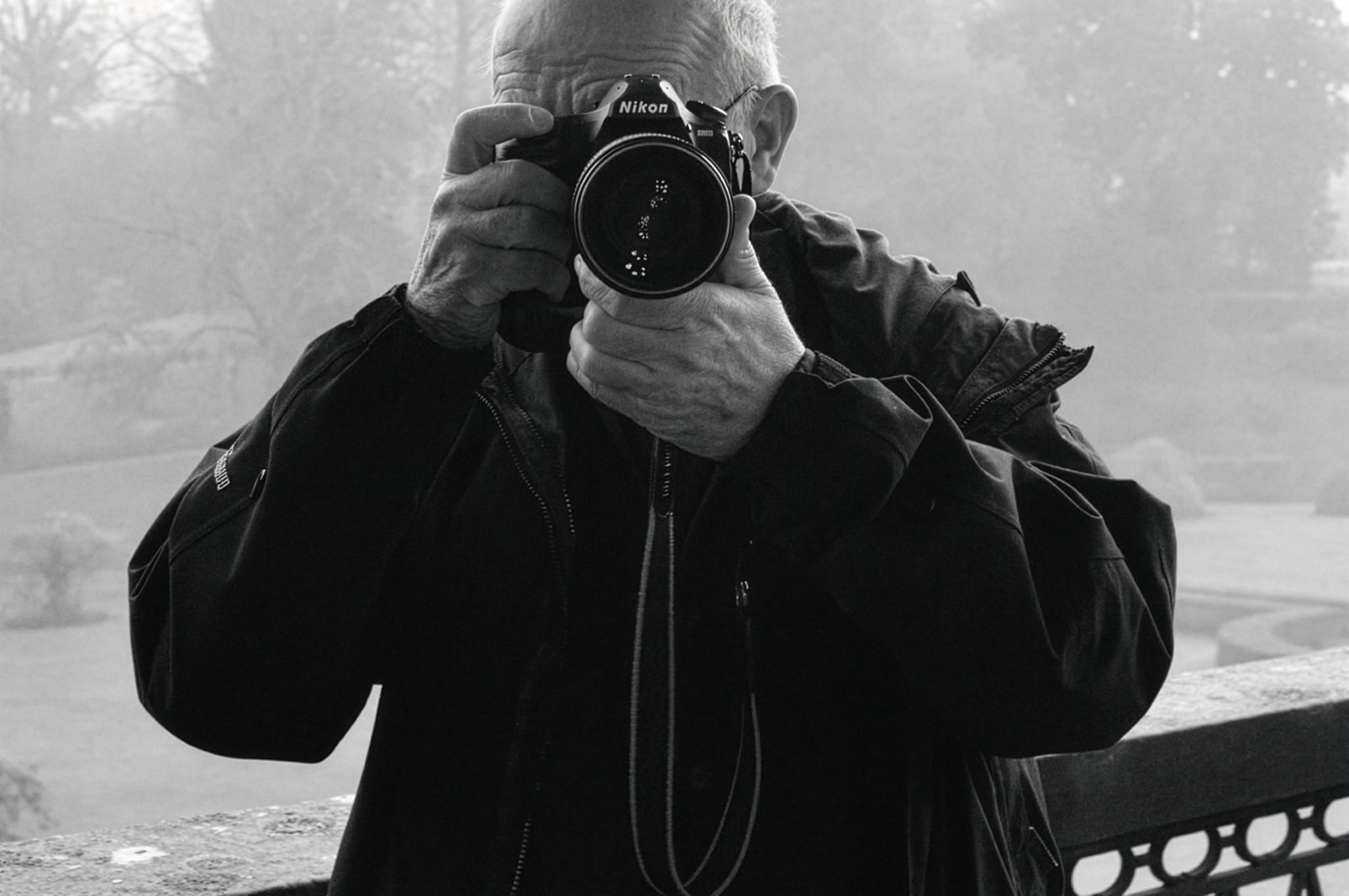 Photographer holding a Nikon camera up to his face, aiming the lens directly at the viewer outdoors