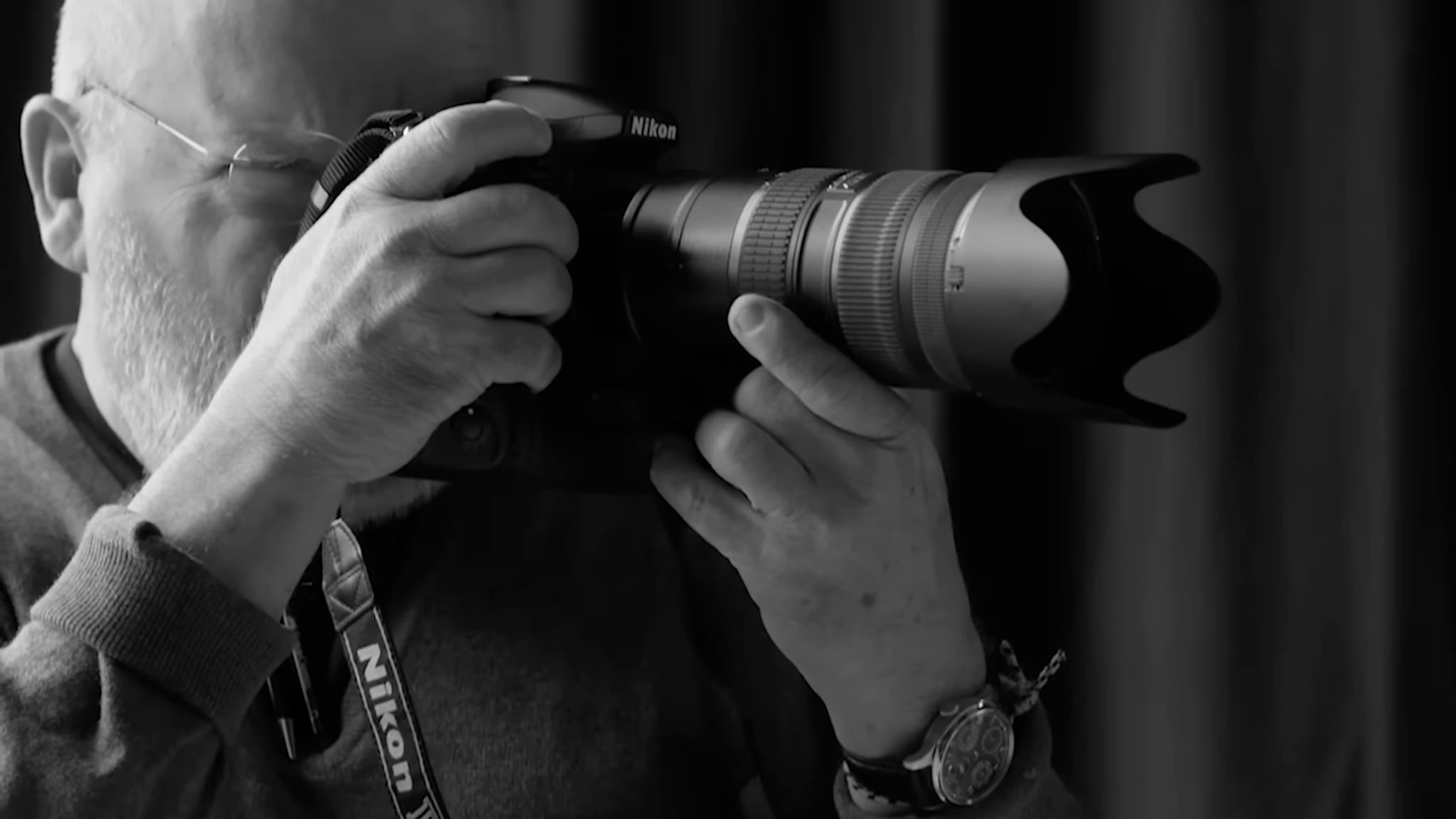 Bearded man with glasses holding a Nikon DSLR camera with a large telephoto lens up to his eye
