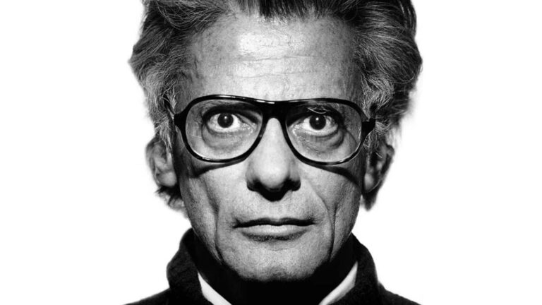 Black and white close-up portrait of an older man with thick dark-rimmed glasses staring directly forward