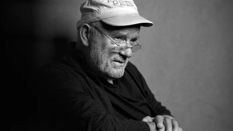 Black and white portrait of an older bearded man wearing eyeglasses and a cap, looking downward thoughtfully
